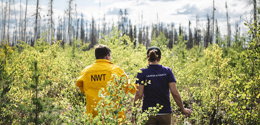 Two people walking through forest wearing NWT and Laurier Research shirts