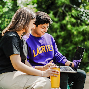 Two students are sitting on a bench, and one showing the other something on their laptop