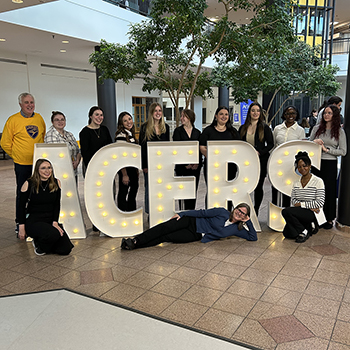 Students and faculty members pose for a photo in front of an illuminated sign reading ACERS